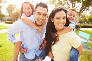 Parents Giving Children Piggyback Ride In Garden Smiling To Camera